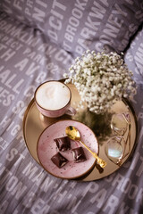 Breakfast set on a bed in pink dishes. Pink cup of cappuccino, pieces of milk chocolate on a pink plate. Vase of white gipsofila. Golden tray with golden glasses.