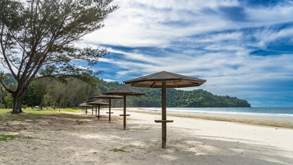 A row of sun umbrellas on the sandy beach. Green grass, trees on the lawn. The waves of the turquoise ocean are foaming. A hill in the distance against the blue sky and clouds. Malaysia. Borneo. 