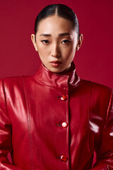 Fashionable asian brunette in a red leather jacket posing with eyes closed against a red background, hands on hips