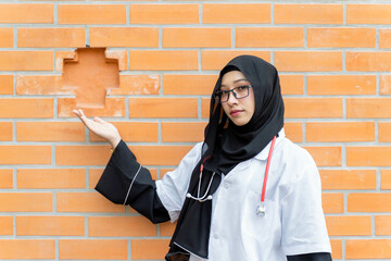 A beautiful Asian Female doctor in hijab traditional clothes and medical shirt portrait on outdoor modern clinic office and brick wall background.