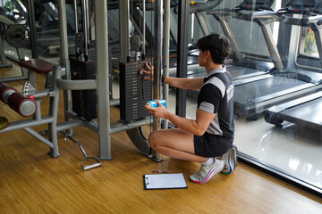 Asian young officer of fitness center checking sling of the weight lifting machine and fitness equipment maintenance list at fitness center.