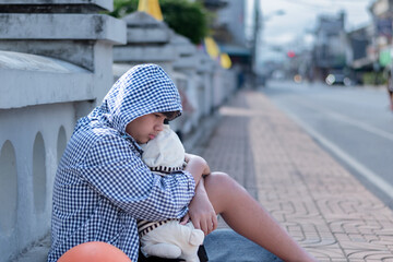 Asian young boy wearing a dark hooded jacket, sitting near a stone fence or barrier thoughtfully...