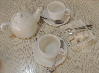 tea party set of white ceramic cups, teapot and sugar bowl with refined sugar and tongs on beige wooden textured table, household items for tea party on tabletop