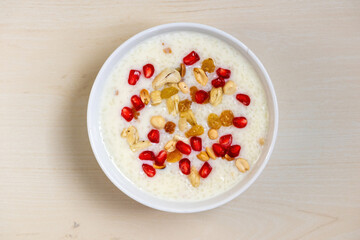 Delicious homemade payesh or payasam, served in a white bowl on a wooden background. Cooked with milk, sugar and rice, it's garnished with pomegranate, cashew nuts, raisins, and peanuts. Top view.