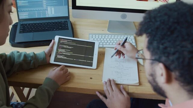 Medium high-angle shot of Middle Eastern male programmer sitting with Chinese female developer at desk at work, looking through computer code on tablet, helping junior colleague fix bugs and errors