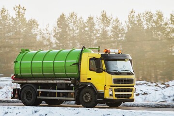 A yellow septic truck with a green tanker is moving on a snowy road amidst trees as the sun sets...