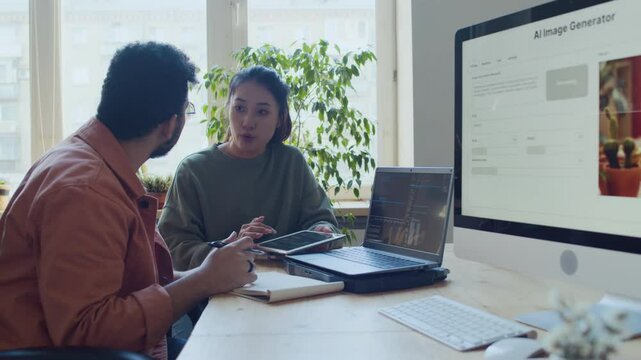 Medium shot of young Asian female and Arab male programmers sitting together at desk, next ot computer with AI generation application on screen, discussing current work and tasks on joint project
