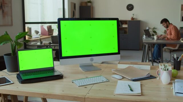 Medium full arc shot of desktop computer and laptop with plain green chromakey screen on desk in innovative IT startup office, male programmer sitting down for work in background. Template, copt space