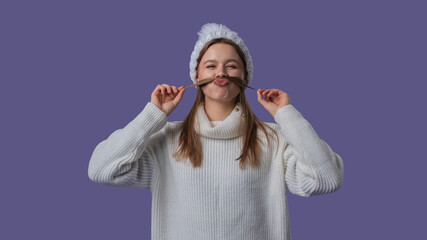 Cheerful young woman in white clothing and knitted hat playfully joking with hair as mustaches,...