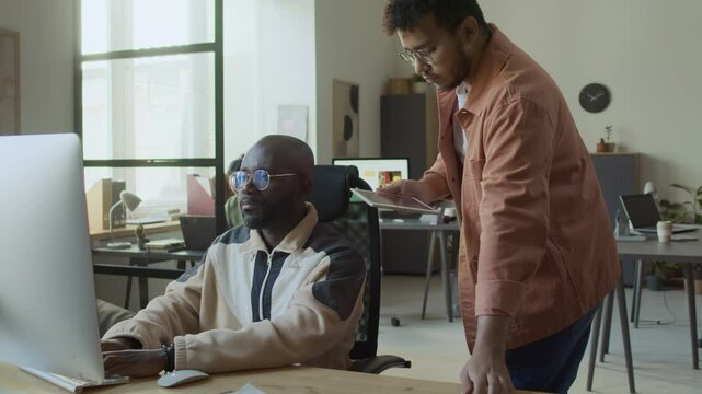 Medium full shot of young Middle Eastern male developer walking up to black colleague typing in computer at desk, while working on joint project, pointing at his code and discussing problem