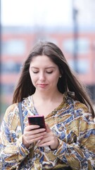 Beautiful woman with long blond hair holding mobile phone, looking at mobile phone screen, using smartphone apps. posing on street. Positive model smiling. She is standing with smartphone in hands on