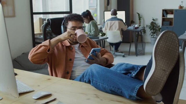 Full shot of cheerful Middle Eastern male programmer in wireless headphones reclining in chair, feet up on desk, enjoying music, using smartphone, drinking tea, while taking break from work