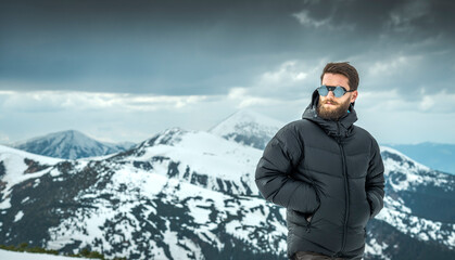 Bearded man wearing black puffer jacket and sunglasses standing in snowy alpine mountains. Cold weather hiking, winter travel and outdoor exploration © XArt
