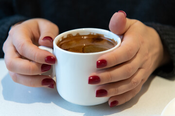Close-up view of female hands with red fingernails holding white blank porcelain cup of americano coffee with brown crema by white table. Soft focus. Copy space. Hot drinks business theme.