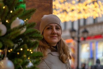 Portrait of serene adult caucasian woman in grey jacket and hat standing by green decorated Christmas tree on city street in a cold cloudy day. Soft focus. People and winter holidays theme.