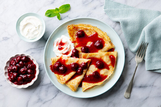 Crepes, thin pancakes with cherry, berry sauce and whipped cream. Marble table background. Top view.