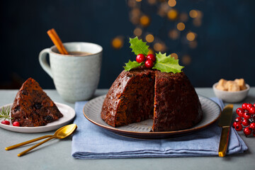 Christmas pudding, fruit cake with mug of tea. Dark background with lights garland. Close up.