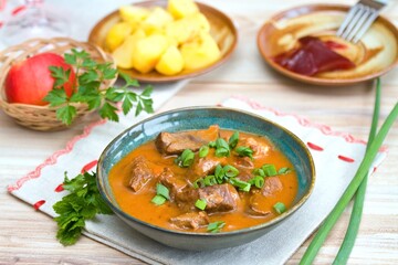 Delicious goulash made from deer meat  with green onion in a bowl. Game stew with potatoes and red currant jam at back.