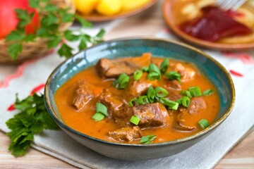 Delicious goulash made from deer meat  with green onion in a bowl. Game stew with potatoes and red currant jam at back.
