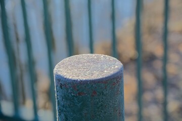 A close-up view of a frost-covered metal fence post with intricate ice crystals, set against a blurred winter background of pale blue and brown hues.