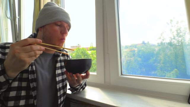A man eats pasta with chopsticks. Eat pasta with wooden chopsticks. Sitting near the window. Slow motion.