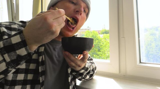 A man eats pasta with chopsticks. Eat pasta with wooden chopsticks. Sitting near the window. Slow motion.