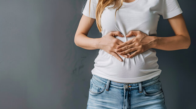 First Weeks Pregnancy Concept: Woman's hands forming a heart shape on her tummy, representing the first weeks of pregnancy, in a white t-shirt and jeans, isolated against a grey background.