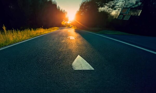 Concept of straight path, direction to success, journey to destination. White arrow on asphalt road during sunset.