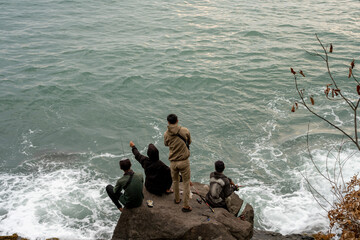 Four people fishing by the rocky seaside, sitting on the rocks as waves crash below. The calm yet...
