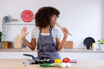 Cheerful young multiracial woman having fun singing using wooden spoon as microphone while...