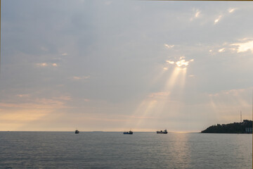 Serene seascape with soft sunlight streaming through clouds, creating beautiful rays over the ocean. Distant boats and a coastal hill add depth and tranquility to the scene.
