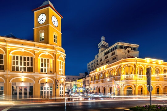Night view of the Phuket Old Town Clock Tower illuminated with vibrant yellow lights, featuring bustling traffic light trails and cultural street decorations.