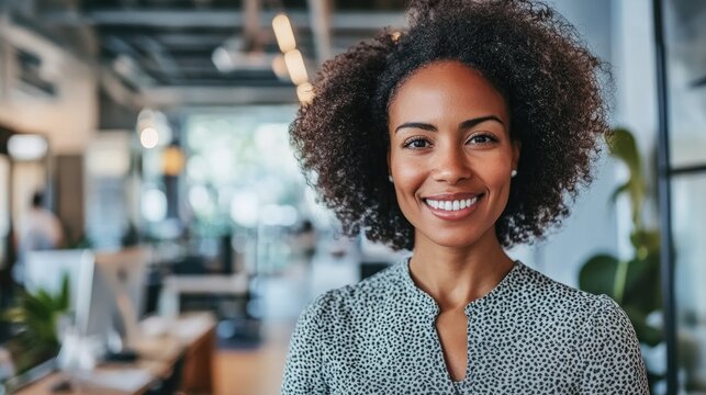 Smiling woman with curly hair in a stylish office, wearing a patterned blouse, exuding confidence and approachability in a vibrant workspace.