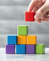 Fototapeta premium A hand places a red block on a colorful pyramid of building blocks, showcasing creativity and architectural play.