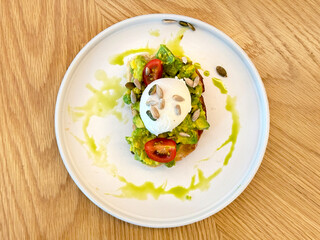 Unique breakfast quiche of avocado bruschetta with poached egg and toasted bread, served on a white plate, isolated on a rustic wooden table.