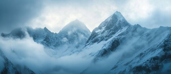 Majestic snow-capped mountains shrouded in mist against a serene cloudy sky at dawn in a breathtaking alpine landscape.