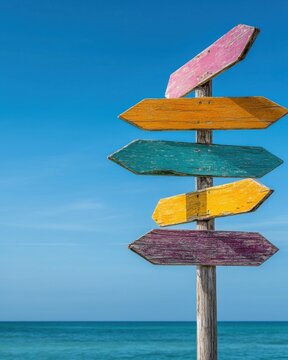 Naklejki A colorful wooden signpost stands against a clear blue sky, pointing in various directions, evoking a sense of adventure by the beach.
