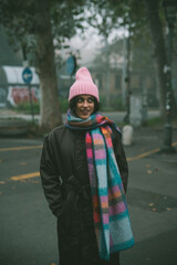 A woman in a pink beanie and colorful scarf stands on a street