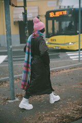 A woman in a pink hat and scarf walks in front of a yellow bus