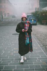 A woman in a pink hat and scarf stands on the sidewalk with a cell phone