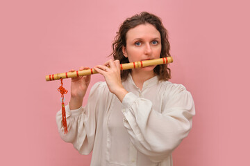 Woman playing bamboo flute decorated with red tassel. Studio portrait on a pink background. Musical performance and traditional culture concept.
