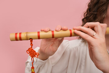 Close-up of a woman playing a bamboo flute with red decorative knot on a pink background. Cultural...