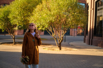 Woman holding autumn leaves and a cup, standing outdoors near trees and buildings on a sunny day. Street scene photography with natural light. Woman 50 years old (fifty year old)