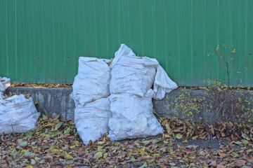 a row of full white garbage bags stand on the ground near a gray green fence wall on a summer street