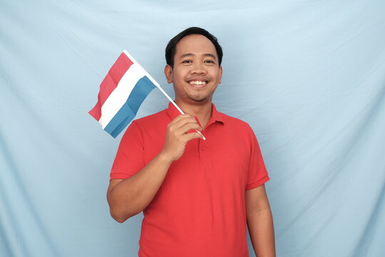 Smiling Indonesian man in a red shirt holding a Netherlands flag, set against a light blue background, showcasing cultural pride.