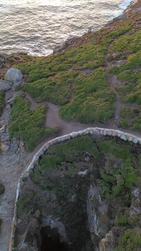 Descent to O Burato do Inferno cave on Ons Island, Galicia. The rocky chasm is bordered by a stone wall and surroundend lush greenery. Behind it, a golden lit cliff stands against calm Atlantic Ocean.