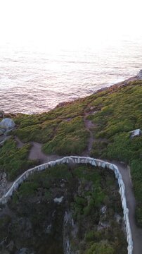 Ascent from O Burato do Inferno cave on Ons Island, Galicia. The rocky chasm is bordered by a stone wall and surroundend lush greenery. Behind it, golden lit cliff stands against calm Atlantic Ocean.