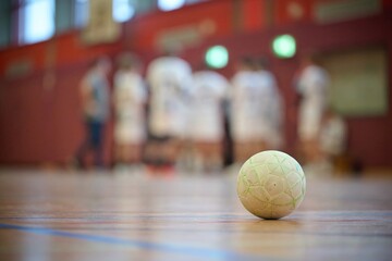 Close-up of a handball on a gym floor with blurred players in the background, depicting a sports...