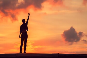 Fototapeta premium Silhouette of a victorious woman standing on a rooftop at sunset, raising her fist in triumph
