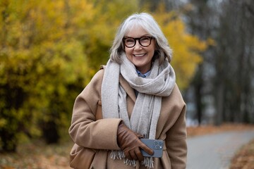 Smiling older woman in a stylish coat and scarf enjoying a walk in a colorful autumn park surrounded by vibrant yellow and orange trees